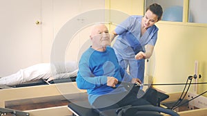 Young female doctor of rehabilitation center helping elderly man to do remedial gymnastic with pilates resistance ring