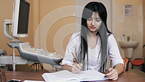 Young female doctor sitting at the desk and writing prescription and filling the forms in clinic, filling out health