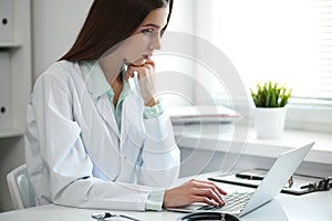 Young female doctor typing on laptop computer while sitting at the table near the window in hospital office