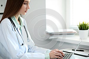 Young female doctor typing on laptop computer while sitting at the table near the window in hospital office