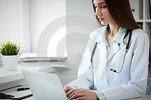 Young female doctor typing on laptop computer while sitting at the table near the window in hospital office