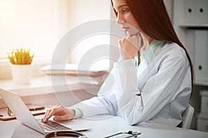 Young female doctor typing on laptop computer while sitting at the table near the window in hospital office