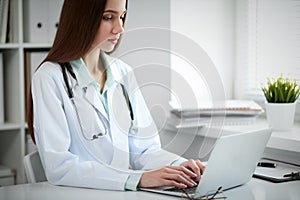 Young female doctor typing on laptop computer while sitting at the table near the window in hospital office