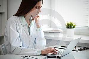 Young female doctor typing on laptop computer while sitting at the table near the window in hospital office