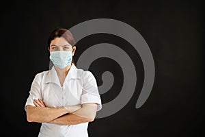 Young female doctor in uniform in a medical mask on a black background. A nurse in a department store folded her arms