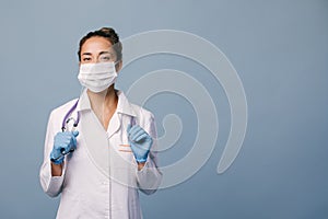 Young female doctor wearing latex gloves and white medical coat holding stethoscope on blue background