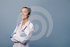 Young female doctor wearing latex gloves and white medical coat holding stethoscope on blue background