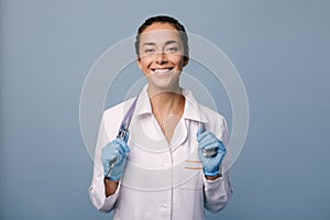 Young female doctor wearing latex gloves and white medical coat holding stethoscope on blue background