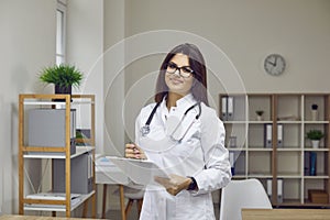 Young female doctor who posing with clipboard in her hands in office of modern medical center.