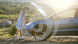 Young female driver standing near a broken car with open up hood inspecting her vehicle engine and waiting for help.