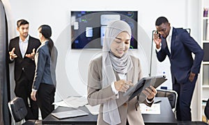 Young female employee stands holding a tablet and is ready for an online meeting for company business. Behind is the atmosphere of