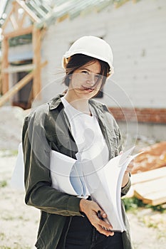 Young female engineer or construction worker in hardhat smiling against of building new modern house. Stylish happy woman