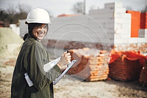 Young female engineer or construction worker in hardhat smiling against of building new modern house. Stylish happy woman