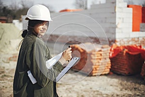 Young female engineer or construction worker in hardhat smiling against of building new modern house. Stylish happy woman