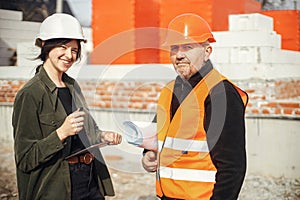 Young female engineer and senior construction worker in hardhat looking at blueprints of new modern house. Stylish woman architect