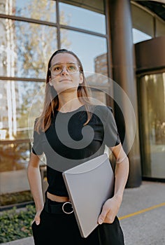 Young female freelancer with laptop standing on modern building background and looks away