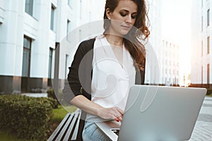 Young female freelancer making labor market research on modern laptop, sits on outdoors in urban street. Woman student work on mob