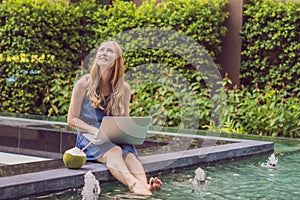 Young female freelancer sitting near the pool with her laptop in the hotel browsing in her smartphone. Busy at holidays