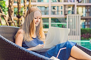 Young female freelancer sitting near the pool with her laptop in the hotel browsing in her smartphone. Busy at holidays