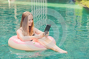 Young female freelancer sitting near the pool with her laptop in the hotel browsing in her smartphone. Busy at holidays