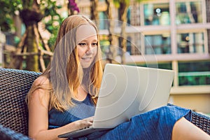 Young female freelancer sitting near the pool with her laptop in the hotel browsing in her smartphone. Busy at holidays. Distant w