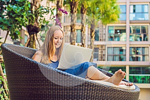 Young female freelancer sitting near the pool with her laptop in the hotel browsing in her smartphone. Busy at holidays. Distant w