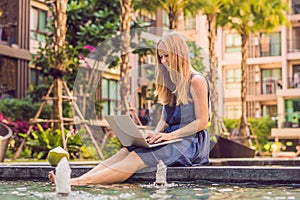 Young female freelancer sitting near the pool with her laptop in the hotel browsing in her smartphone. Busy at holidays. Distant w