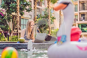 Young female freelancer sitting near the pool with her laptop in the hotel browsing in her smartphone. Busy at holidays. Distant w