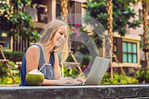 Young female freelancer sitting near the pool with her laptop in the hotel browsing in her smartphone. Busy at holidays. Distant w