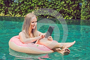 Young female freelancer sitting near the pool with her laptop in the hotel browsing in her smartphone. Busy at holidays. Distant w