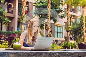 Young female freelancer sitting near the pool with her laptop in the hotel browsing in her smartphone. Busy at holidays. Distant w