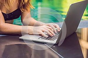 Young female freelancer sitting near the pool with her laptop in the hotel browsing in her smartphone. Busy at holidays. Distant w