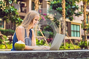 Young female freelancer sitting near the pool with her laptop in the hotel browsing in her smartphone. Busy at holidays. Distant w