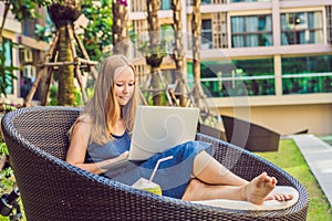 Young female freelancer sitting near the pool with her laptop in the hotel browsing in her smartphone. Busy at holidays. Distant w