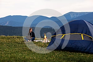 Young female freelancer working on laptop in the mountains in the evening. Tourist girl sitting near campfire and tent. Copy space