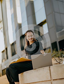 Young female freelancer working on laptop while sitting on modern building background