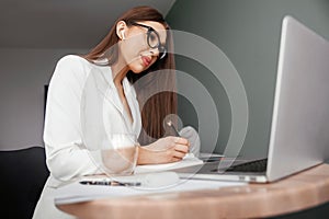 Young female in glasses using laptop, communicates on internet with customer in home, coffee mug on table.Cozy office workplace