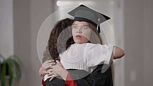 Young female graduate in gown and hat hugging woman with dissatisfied irritated facial expression. Caucasian