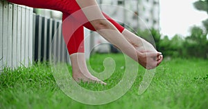 Young female gymnast woman warms her leg before training on the green grass outdoors, fitness in the urban environment