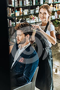 Young female hairdresser smiling at camera while drying hair to handsome client