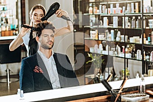 Young female hairstylist combing and drying hair to handsome client