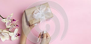 Young female hands hold a gift craft box with a white bow on a pink background with orchid flowers