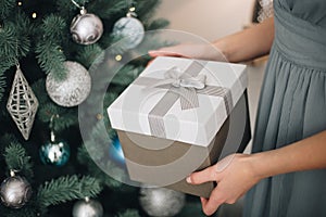 Young female hands holding gift box near the Christmas tree. New Year atmosphere