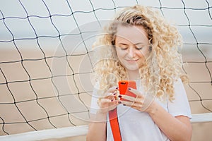 Young female has curly hair holds modern smart phone, downloads photos from social networks, poses against tennis net background,
