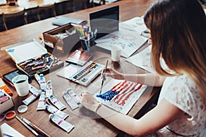 Young female illustrator drawing flag of the USA in using aquarelle paint sitting at workplace