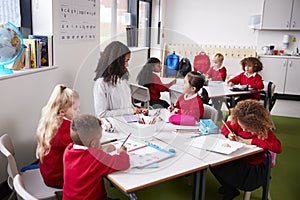Young female infant school teacher sitting at a table in a classroom with her pupils
