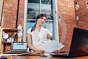 Young female journalist preparing a new article holding papers using laptop sitting at desk in creative office