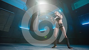 A young female kickboxer kicking a black punching bag with a powerful kick. Athlete practicing attack exercises in a