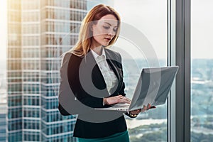 Young female lawyer working in her luxurious office holding a laptop standing against panoramic window with a view on