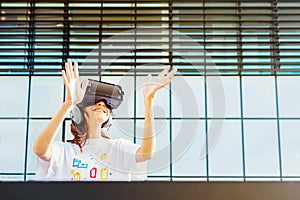 Young female with long hair and white t-shirt wearing virtual reality glasses and big headphones while raising hands in front of